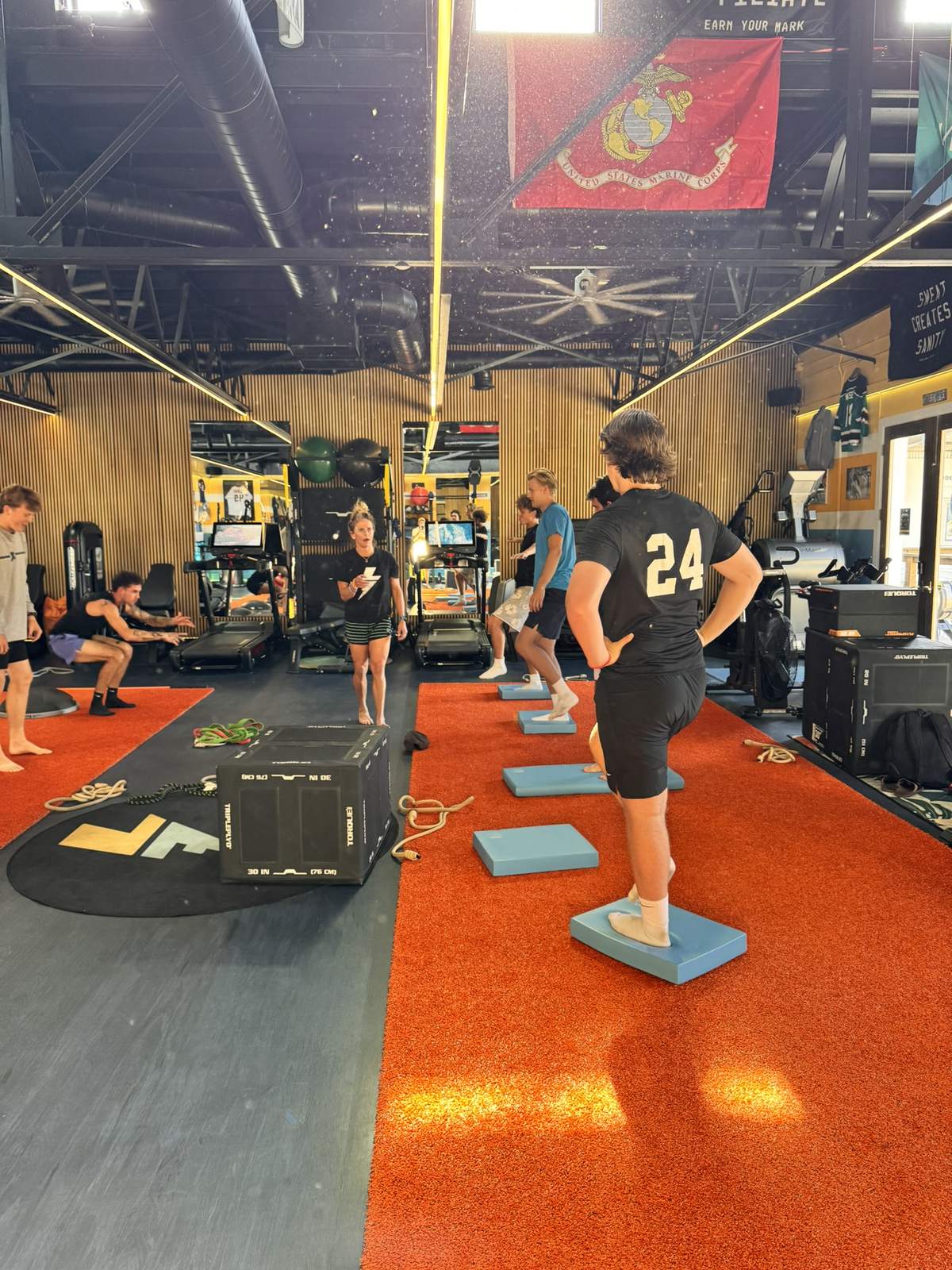 A class in motion at Lilley Fitness: young athletes balance on blue pads on the orange turf while Erika coaches in the background.